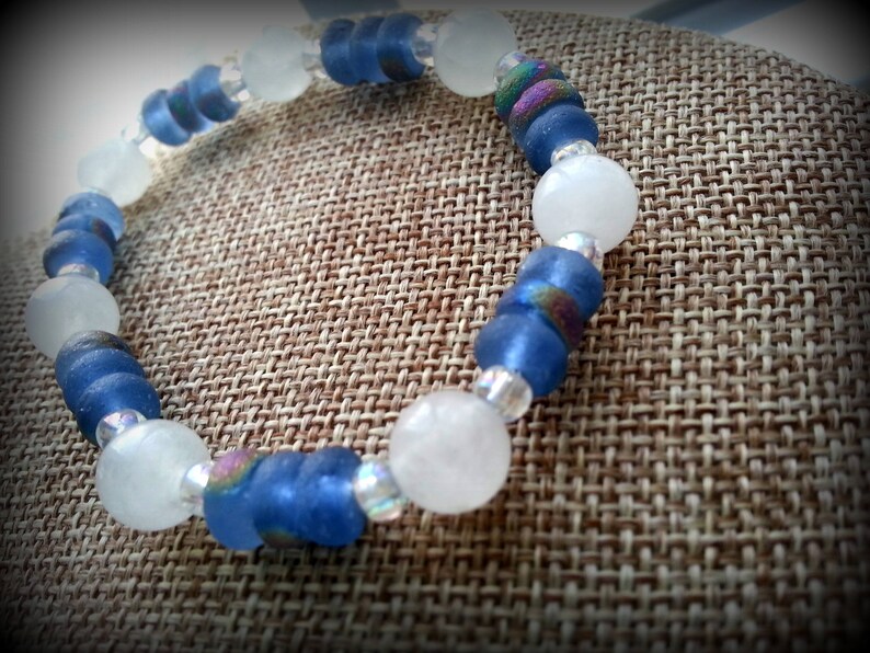May include: A blue and white beaded bracelet with iridescent accents. The bracelet is made of glass beads and is displayed on a brown fabric background.