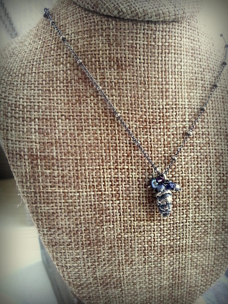 May include: A silver chain necklace with a small, detailed pinecone charm and purple beads.
