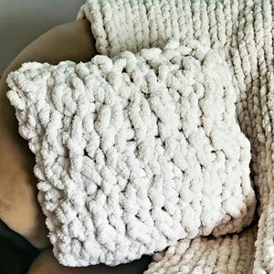 May include: A cream-colored, chunky knit pillow and blanket set. The pillow is square and rests on a matching blanket. The thick yarn creates a textured, cozy appearance. The set is likely for home decor and comfort.