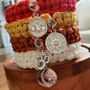 May include: Four colorful crocheted bracelets with silver charms. The charms include an eye with a crystal, a star with a crystal, an Om symbol, and a crescent moon with crystals.