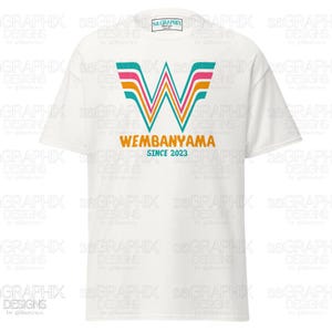 May include: White t-shirt featuring a colorful graphic design. The design includes a stylized "W" with pink, orange, and teal accents, along with the text "WEMBANYAMA SINCE 2023" in orange. The shirt has a classic crew neck.