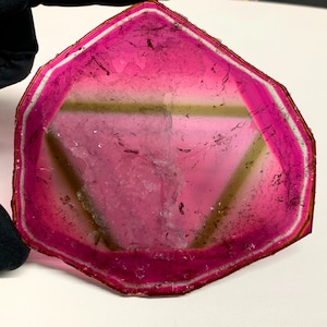 K&ouml;nnte beinhalten: Eine durchscheinende, rosa Edelstein-Scheibe mit wei&szlig;em und braunem Rand. Der Stein hat eine dreieckige Form mit einem Farbverlauf, der von einem tiefen Rosa zu einem helleren Farbton in der Mitte &uuml;bergeht. Die Kanten sind facettiert.