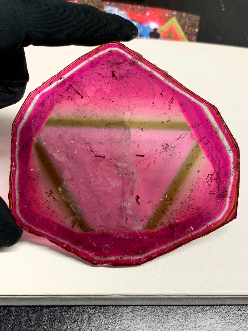 K&ouml;nnte beinhalten: Eine durchscheinende, rosa Edelstein-Scheibe mit wei&szlig;em und braunem Rand. Der Stein hat eine dreieckige Form mit einem Farbverlauf, der von einem tiefen Rosa zu einem helleren Farbton in der Mitte &uuml;bergeht. Die Kanten sind facettiert.