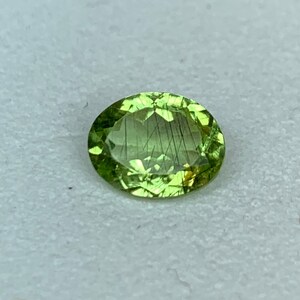 May include: Oval-cut, light green gemstone with internal black inclusions. The gem has a faceted surface, reflecting light. The stone is set against a neutral background, highlighting its color and clarity. This gemstone is suitable for jewelry making.