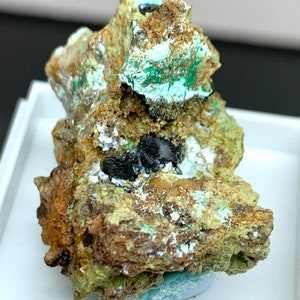 May include: A close-up of a mineral specimen featuring a rough, textured surface. The rock displays a mix of colors, including brown, green, and black. The mineral has a crystalline structure with a few dark, shiny spots.