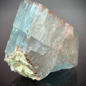 Puede incluir: Una formaci&oacute;n cristalina transl&uacute;cida de color azul claro con inclusiones de color marr&oacute;n rojizo y un grupo de peque&ntilde;os cristales blanquecinos en la base. La superficie del cristal es rugosa y texturizada.