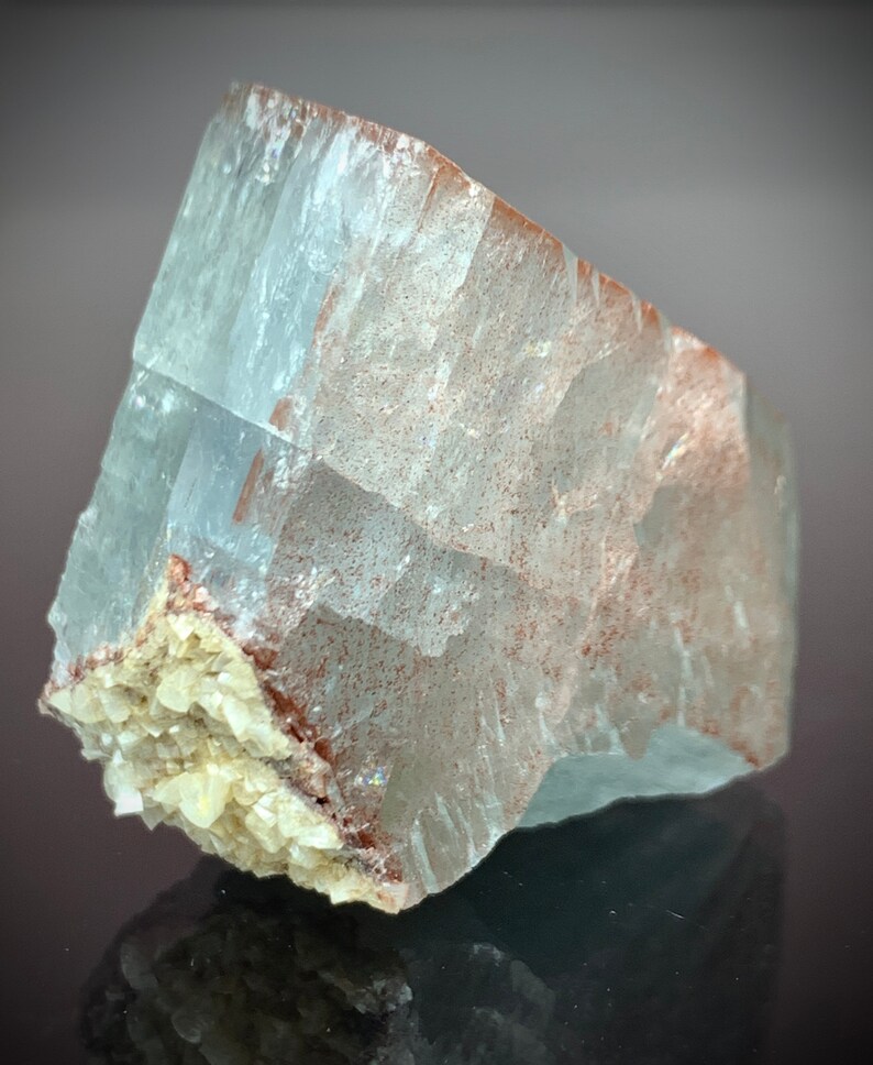 Puede incluir: Una formaci&oacute;n cristalina transl&uacute;cida de color azul claro con inclusiones de color marr&oacute;n rojizo y un grupo de peque&ntilde;os cristales blanquecinos en la base. La superficie del cristal es rugosa y texturizada.