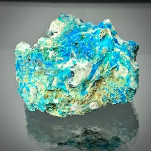 May include: A close-up of a vibrant mineral specimen featuring a striking combination of colors. The rock displays a rich turquoise hue, interspersed with patches of bright blue and hints of green. The surface has a rough, textured appearance, with areas of white and brown.