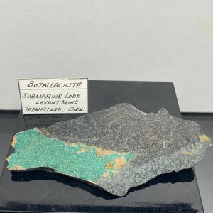 May include: A Botallackite mineral specimen from the Submarine Lode, Levant Mine, Trewellard, Cornwall. The rock is dark grey with a patch of vibrant turquoise-green and hints of orange.