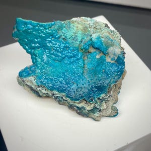 May include: A vibrant, textured mineral specimen with a striking turquoise hue. The surface displays a bubbly, almost coral-like texture, contrasted by areas of darker blue and greyish-white. The mineral sits on a white surface, showcasing its natural beauty.