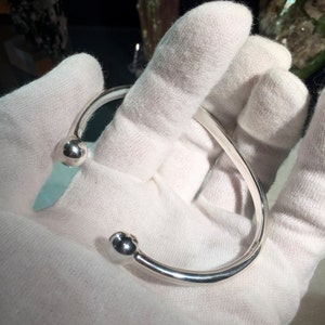 May include: A silver open cuff bracelet with a curved design. Each end of the bracelet has a small, round ball. The bracelet is held in a gloved hand, highlighting its polished finish and simple elegance. The bracelet is a piece of jewellery.
