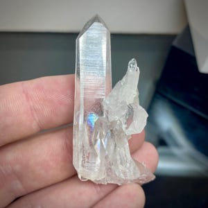 May include: A clear quartz crystal cluster with a large, pointed main crystal and several smaller crystals attached. The quartz has a polished, reflective surface with iridescent rainbow flashes. The crystal is held in a hand.