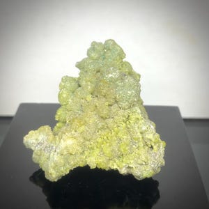 May include: A close-up of a mineral specimen with a textured, crystalline structure. The mineral is primarily a vibrant yellow-green color, with some areas appearing lighter. The specimen is displayed on a black surface.