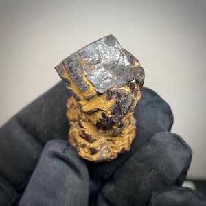 Può includere: Primo piano di un campione minerale con una parte superiore grigio scuro metallico e una base marrone dorato strutturata. Il minerale ha un aspetto stratificato, con strutture cristalline visibili. Il campione è tenuto in una mano guantata nera.
