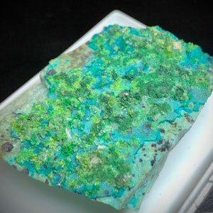 Può includere: Primo piano di un campione minerale con un mix di colori. La roccia ha una base turchese e verde acqua, con macchie di verde brillante e punti scuri. La trama appare ruvida e irregolare, con formazioni cristalline.