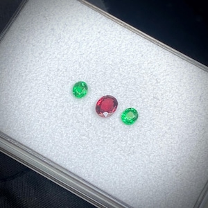 May include: Three gemstones: two oval-cut emeralds and a ruby. The emeralds are a vibrant green, while the ruby is a deep red. The gems are displayed on a white surface inside a clear case.