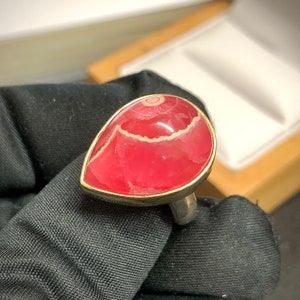 Puede incluir: Un anillo en forma de lágrima con una vibrante piedra preciosa roja con bandas blancas y rosas. La piedra está engastada en un bisel dorado y montada sobre una banda plateada. El anillo se sostiene sobre un fondo de tela negra.