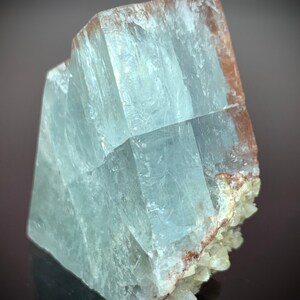 Puede incluir: Formaci&oacute;n cristalina transl&uacute;cida de color azul claro con forma rectangular. El cristal tiene una apariencia en capas con estructuras internas visibles. Los bordes tienen un tono marr&oacute;n rojizo y la base presenta peque&ntilde;os grupos cristalinos blanquecinos.