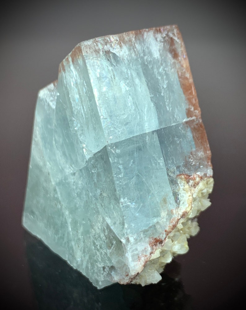 Puede incluir: Formaci&oacute;n cristalina transl&uacute;cida de color azul claro con forma rectangular. El cristal tiene una apariencia en capas con estructuras internas visibles. Los bordes tienen un tono marr&oacute;n rojizo y la base presenta peque&ntilde;os grupos cristalinos blanquecinos.