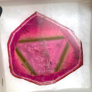 K&ouml;nnte beinhalten: Eine lebendige, rosa und klare Edelsteinscheibe mit einem geometrischen Design. Der Stein hat einen dunkelrosa Rand und eine hellere rosa Mitte, mit einem sichtbaren dreieckigen Muster. Der Edelstein wird in einer wei&szlig;en Box pr&auml;sentiert.