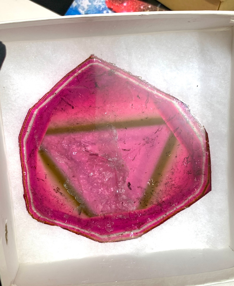 K&ouml;nnte beinhalten: Eine lebendige, rosa und klare Edelsteinscheibe mit einem geometrischen Design. Der Stein hat einen dunkelrosa Rand und eine hellere rosa Mitte, mit einem sichtbaren dreieckigen Muster. Der Edelstein wird in einer wei&szlig;en Box pr&auml;sentiert.