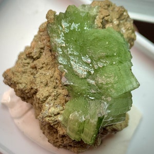 May include: A close-up of a mineral specimen featuring a vibrant, translucent green crystal formation. The crystal is layered and sits atop a rough, tan-colored matrix. The specimen is displayed on a white surface.