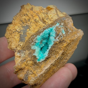May include: A rough, tan-colored rock with a cavity filled with vibrant, crystalline turquoise formations. The rock's surface has a textured appearance with darker brown and gray areas, creating a striking contrast with the bright blue-green crystals.