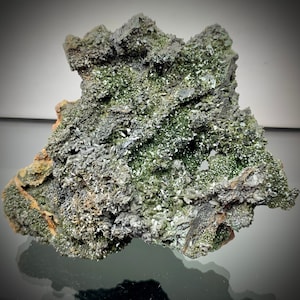 May include: A close-up of a mineral specimen featuring a complex arrangement of dark gray and green crystals. The surface has a rough, textured appearance with areas of sparkling, light-colored crystalline formations. The overall shape is irregular and jagged.