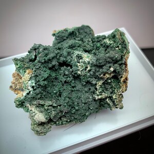 Puede incluir: Primer plano de un espécimen mineral con una superficie texturizada de color verde oscuro y apariencia burbujeante. El mineral tiene una forma rugosa e irregular con áreas de tonos verdes y beige más claros.