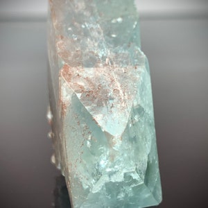 Puede incluir: Cristal de aguamarina azul claro transl&uacute;cido, de forma rectangular. El cristal tiene un aspecto facetado con estructuras internas visibles y algunas inclusiones de color marr&oacute;n rojizo. La est&eacute;tica general es natural y cristalina.