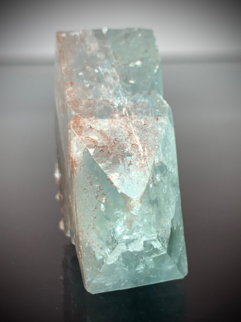 Puede incluir: Cristal de aguamarina azul claro transl&uacute;cido, de forma rectangular. El cristal tiene un aspecto facetado con estructuras internas visibles y algunas inclusiones de color marr&oacute;n rojizo. La est&eacute;tica general es natural y cristalina.