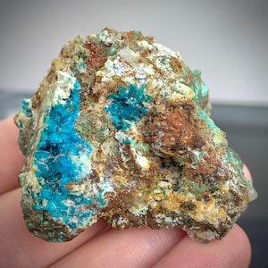 May include: A close-up of a mineral specimen featuring a mix of colors. The rock has a rough, textured surface with areas of vibrant turquoise blue, rusty brown, and patches of white and green. The mineral appears to be a natural formation.