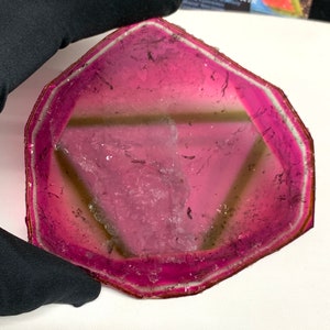 K&ouml;nnte beinhalten: Eine durchscheinende, rosa und hellbraune Edelsteinscheibe mit geometrischer Form. Der Stein hat einen leuchtend rosa Rand, der in eine hellere hellbraune Mitte &uuml;bergeht. Die Kanten sind mit einer d&uuml;nnen, silberfarbenen Zierleiste umrandet.