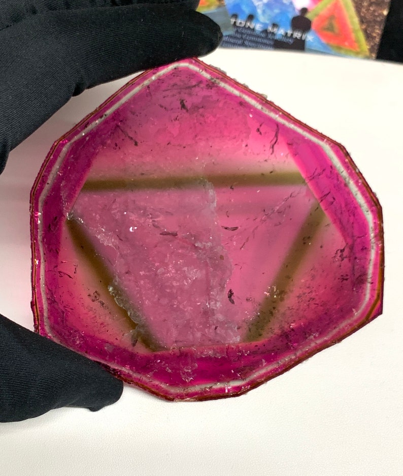 K&ouml;nnte beinhalten: Eine durchscheinende, rosa und hellbraune Edelsteinscheibe mit geometrischer Form. Der Stein hat einen leuchtend rosa Rand, der in eine hellere hellbraune Mitte &uuml;bergeht. Die Kanten sind mit einer d&uuml;nnen, silberfarbenen Zierleiste umrandet.