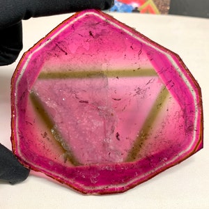K&ouml;nnte beinhalten: Ein facettierter Turmalin-Edelstein in Wassermelonenfarben. Der Edelstein hat einen leuchtend rosa &auml;u&szlig;eren Rand, der in eine hellgr&uuml;ne Mitte &uuml;bergeht. Der Stein wird von einer behandschuhten Hand gehalten, die seine nat&uuml;rliche Sch&ouml;nheit und den einzigartigen Farbverlauf hervorhebt.