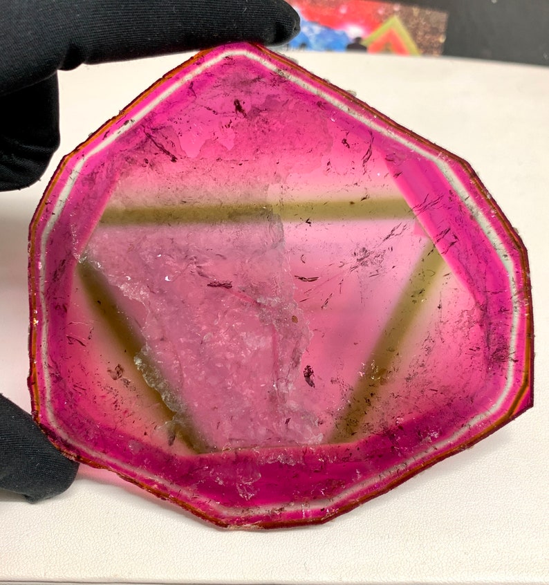 K&ouml;nnte beinhalten: Ein facettierter Turmalin-Edelstein in Wassermelonenfarben. Der Edelstein hat einen leuchtend rosa &auml;u&szlig;eren Rand, der in eine hellgr&uuml;ne Mitte &uuml;bergeht. Der Stein wird von einer behandschuhten Hand gehalten, die seine nat&uuml;rliche Sch&ouml;nheit und den einzigartigen Farbverlauf hervorhebt.