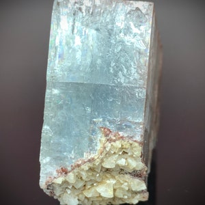 Puede incluir: Una formaci&oacute;n cristalina rectangular y transl&uacute;cida, de color azul claro. El cristal tiene una superficie rugosa y texturizada, y un grupo de peque&ntilde;os cristales beige en la base. El cristal se muestra sobre un fondo oscuro.
