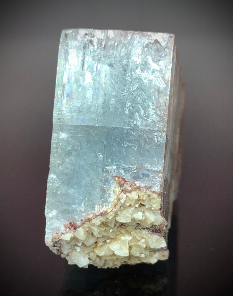 Puede incluir: Una formaci&oacute;n cristalina rectangular y transl&uacute;cida, de color azul claro. El cristal tiene una superficie rugosa y texturizada, y un grupo de peque&ntilde;os cristales beige en la base. El cristal se muestra sobre un fondo oscuro.