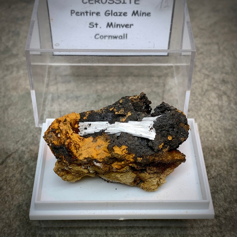 May include: A mineral specimen of Cerussite from the Pentire Glaze Mine in St. Minver, Cornwall, presented in a clear display case. The mineral features a dark, textured matrix with white, crystalline formations and areas of orange and brown.