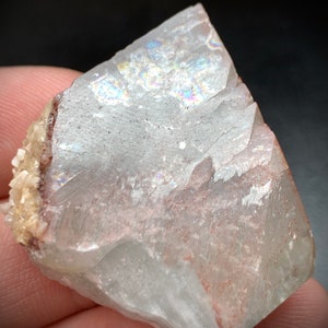 Puede incluir: Un esp&eacute;cimen mineral transl&uacute;cido con &aacute;reas iridiscentes. La piedra tiene una superficie rugosa y fracturada con tonos rojos y beige. El mineral se sostiene en una mano sobre un fondo oscuro.