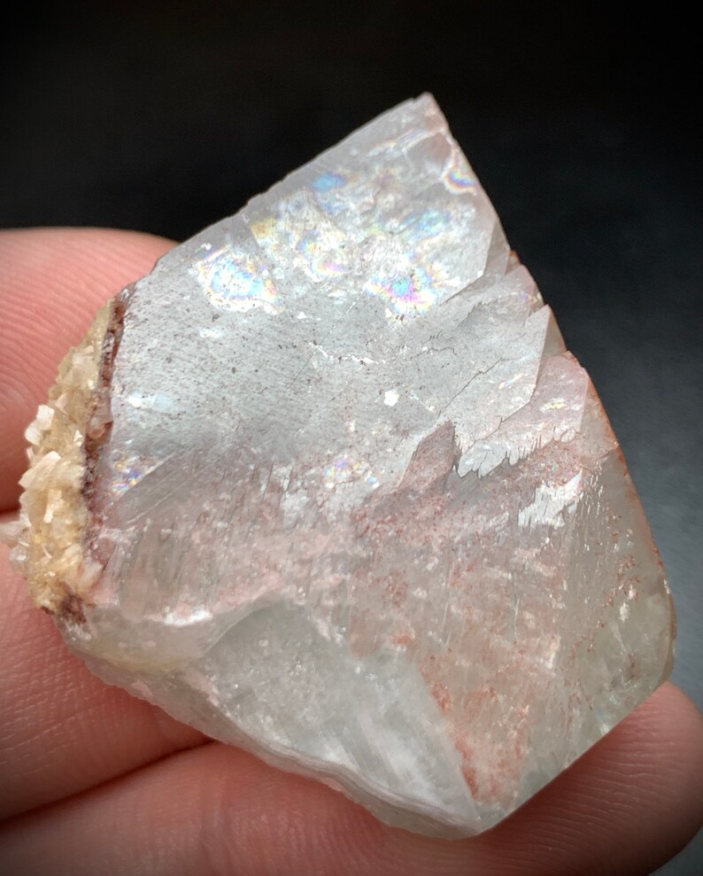 Puede incluir: Un esp&eacute;cimen mineral transl&uacute;cido con &aacute;reas iridiscentes. La piedra tiene una superficie rugosa y fracturada con tonos rojos y beige. El mineral se sostiene en una mano sobre un fondo oscuro.