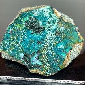 Può includere: Un vivace esemplare minerale di forma irregolare con una sorprendente superficie verde e turchese. La pietra presenta formazioni scure e appuntite e piccole macchie bianche, su una matrice ruvida grigio-marrone. La superficie del minerale ha un aspetto strutturato.