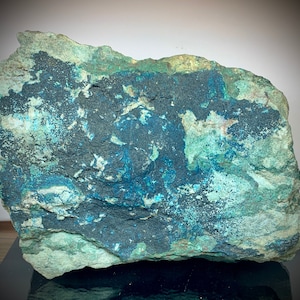 May include: A large, rough-hewn rock formation with a mix of colors. The stone features areas of deep blue, teal, and black, contrasted against a lighter, greenish-gray base. The texture appears coarse and natural, suggesting a mineral specimen.