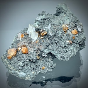 May include: A close-up of a mineral specimen featuring a dark gray matrix with embedded crystals. The crystals are a mix of clear, white, and golden-brown hues. The specimen has a rough, textured surface and is displayed against a dark background.
