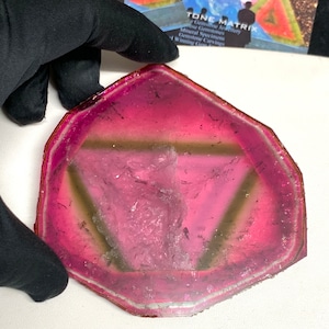 K&ouml;nnte beinhalten: Eine transluzente, unregelm&auml;&szlig;ig geformte Edelsteinscheibe mit einem lebendigen Farbverlauf von Rosa und Magenta. Der Stein hat eine hellere, fast klare Mitte mit einem dunkleren rosa Rand. Ein dreieckiges Muster ist im Stein sichtbar. Der Edelstein wird von einer behandschuhten Hand gehalten.