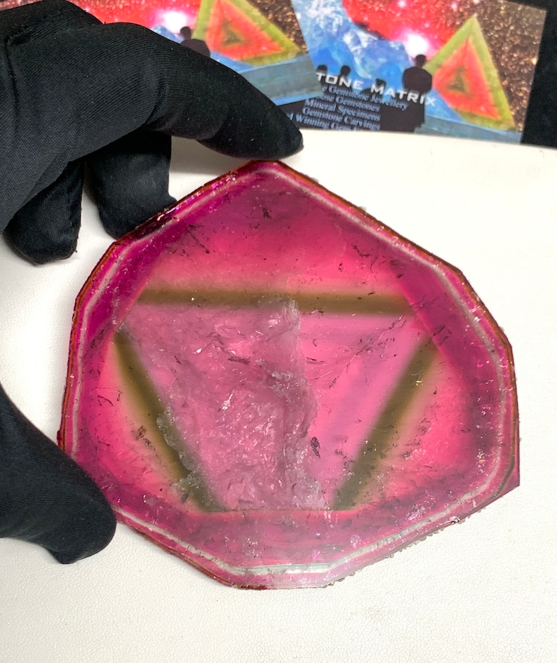K&ouml;nnte beinhalten: Eine transluzente, unregelm&auml;&szlig;ig geformte Edelsteinscheibe mit einem lebendigen Farbverlauf von Rosa und Magenta. Der Stein hat eine hellere, fast klare Mitte mit einem dunkleren rosa Rand. Ein dreieckiges Muster ist im Stein sichtbar. Der Edelstein wird von einer behandschuhten Hand gehalten.