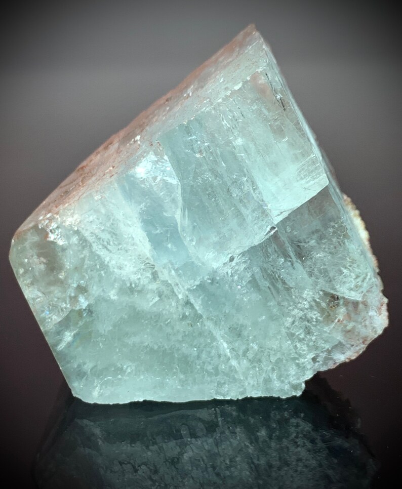 Puede incluir: Un gran cristal de aguamarina transl&uacute;cido con forma rectangular. El cristal tiene un tono azul verdoso p&aacute;lido y una superficie rugosa y texturizada. Los bordes son afilados y bien definidos, y el cristal parece estar parcialmente pulido.