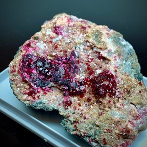 May include: A natural mineral specimen featuring a matrix of tan and gray rock with vibrant, deep red crystalline formations. The crystals are clustered in pockets, creating a striking contrast against the neutral background.