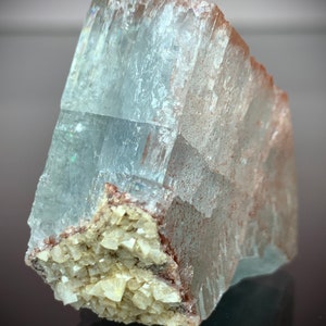 Puede incluir: Una formaci&oacute;n cristalina transl&uacute;cida de color azul claro con una textura rugosa y en capas. El cristal presenta un grupo de peque&ntilde;as estructuras cristalinas blanquecinas en su base, con inclusiones de color marr&oacute;n rojizo. La superficie refleja la luz, creando un efecto brillante.