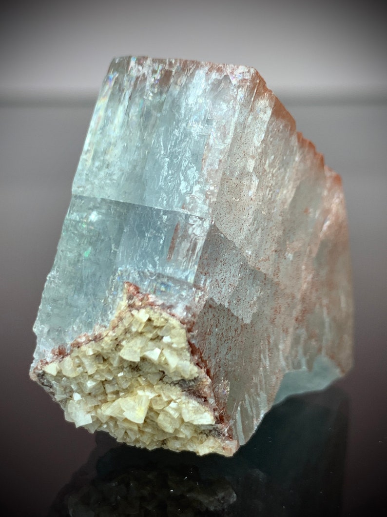 Puede incluir: Una formaci&oacute;n cristalina transl&uacute;cida de color azul claro con una textura rugosa y en capas. El cristal presenta un grupo de peque&ntilde;as estructuras cristalinas blanquecinas en su base, con inclusiones de color marr&oacute;n rojizo. La superficie refleja la luz, creando un efecto brillante.